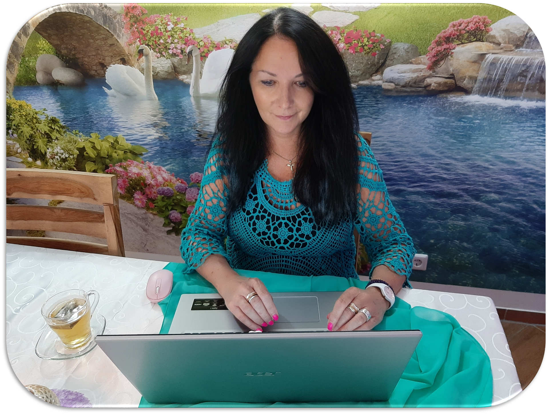 Frau mit langem schwarzem Haar sitzt am Laptop am Tisch, im Hintergrund Wasserfall und Schwäne.